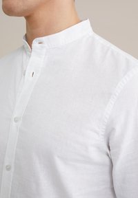 Vit linneskjorta med mandarin-krage, placket med en knapp och subtil textur. Knapparna är rundade och tillverkade av klart plast.