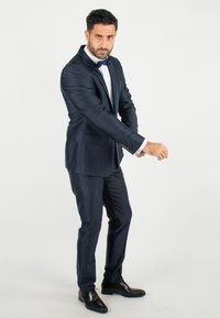 Costume bleu marine avec une coupe ajustée, comprenant une veste à simple boutonnage, un pantalon assorti et un nœud papillon noir. Chaussures en cuir noir.