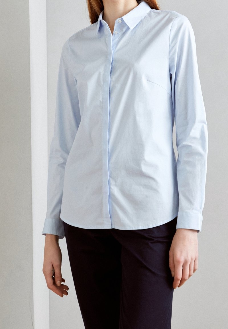 Femme portant une chemise bleu clair à manches longues boutonnée et un pantalon noir, debout devant un fond uni clair.