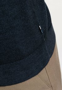 Navyblauer Strickpullover mit geripptem Saum und Seitennähten, abgebildet neben einem sandfarbenen Stoff, der Textur und Farbkontrast hervorhebt.