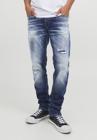 Jasnoniebieskie, postrzępione jeansy z licznymi przetarciami i wyblaknięciami; dopasowany fason, pięć kieszeni, zestawione z czarnymi trampkami z białymi sznurówkami.