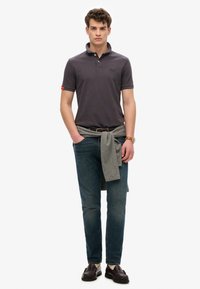 Mørkegrå polo-shirt med krave, blå jeans og en grå sweater bundet om taljen. Brune loafers og et ur fuldender outfittet.