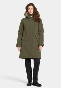 Oliven grøn parka med en faux fur-foring i hætten, lynlås foran, to sidelommer og knælangt design. Sorte bukser og støvler fuldender looket.