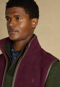 Bordový fleecový vest s šedými akcentmi, so zipsom, vrstvený cez tmavozelenú tweedovú košeľu a modrý šál s vzorom.