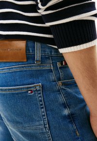 Vaqueros de mezclilla azul con parche del logo de Tommy Hilfiger y pequeño detalle de bandera, combinados con un suéter de punto a rayas azul marino y blanco con la manga visible.