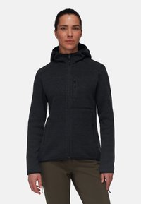 Black zip-up fleece jacket with a hood, textured fabric, side and chest pockets, and fitted design. Worn with khaki pants.