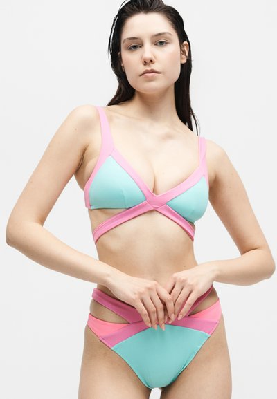 Νέα γυναίκα που φοράει μπλε και ροζ δίπλο μαγιό με λεπτομέρειες κοψίματος, στέκεται με τα χέρια ενωμένα σε λευκό φόντο.