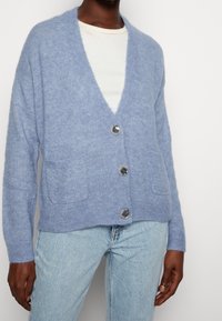 Hellblauer, weicher Strickcardigan mit tiefem V-Ausschnitt, vier Knöpfen, zwei Vorderseitentaschen und gerippten Bündchen.