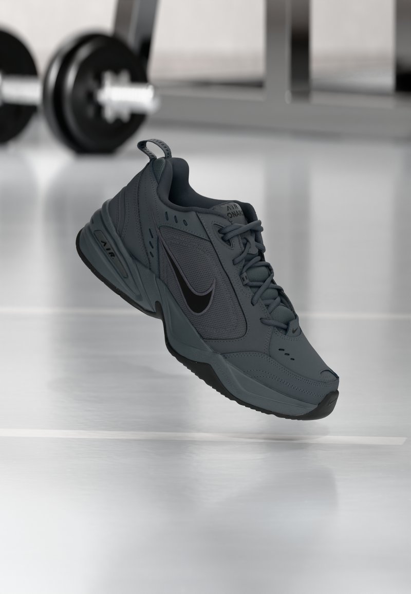 Sapatilha Nike Air Monarch cinza escura suspensa no interior, com pesos de ginásio desfocados ao fundo sobre um piso brilhante.