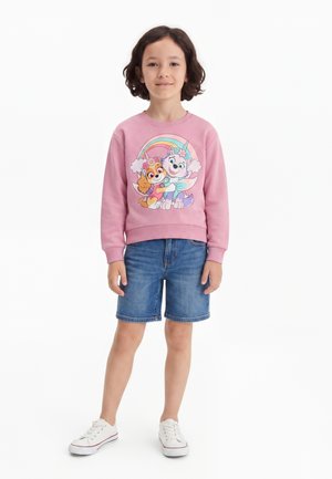 Enfant aux cheveux courts et foncés portant un sweat-shirt rose avec des chiens dessinés et un arc-en-ciel, un short en jean bleu et des baskets blanches, debout sur fond blanc.