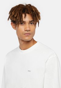 Weißes Langarm-T-Shirt aus Baumwolle. Mit Rundhalsausschnitt und kleinem blauen Dickies-Logo-Patch auf der linken Brust. Glatte Textur.