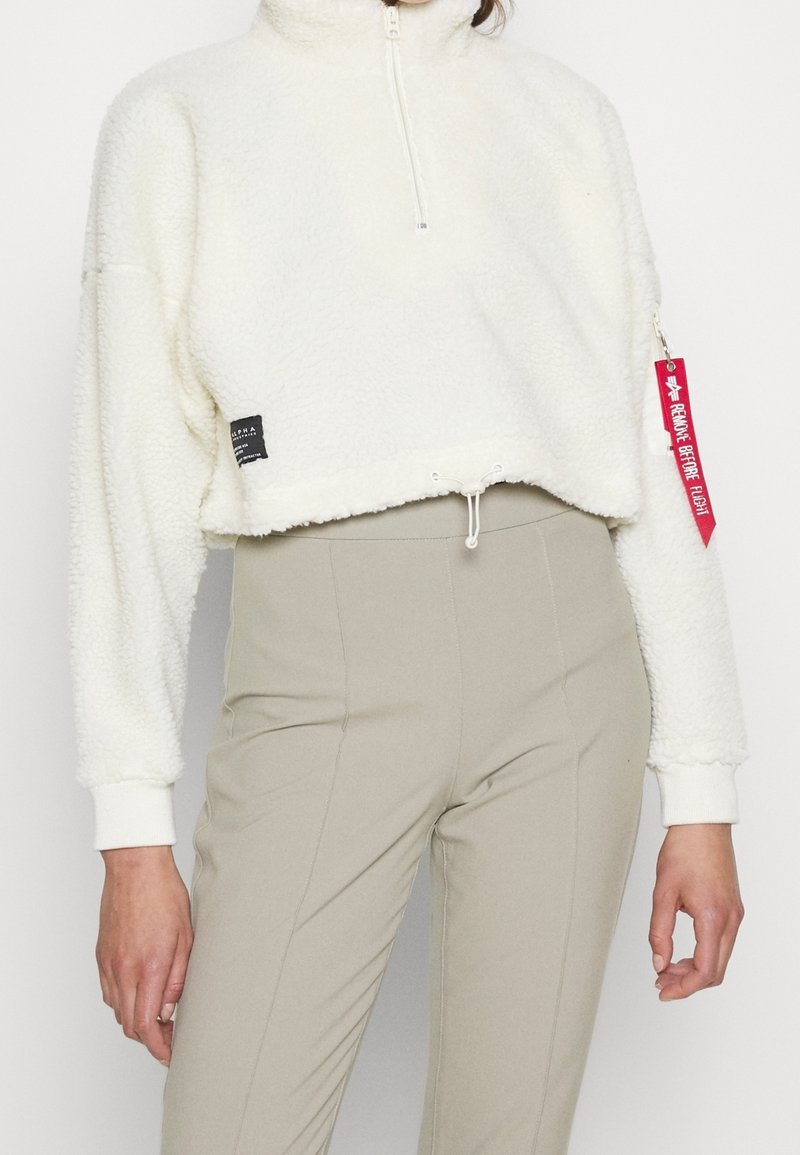 Pull polaire blanc court avec col montant et fermeture éclair quart de longueur. Comprend une étiquette en tissu sur le côté et une boucle rouge sur la manche.
