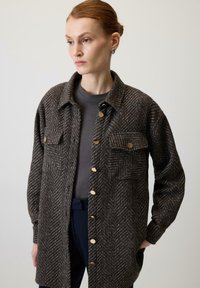 Brązowo-szara koszula typu shirt jacket o fakturze jodełki, z dwoma kieszeniami na piersi, złotymi guzikami i luźnym krojem, noszona na ciemnoszarym topie.