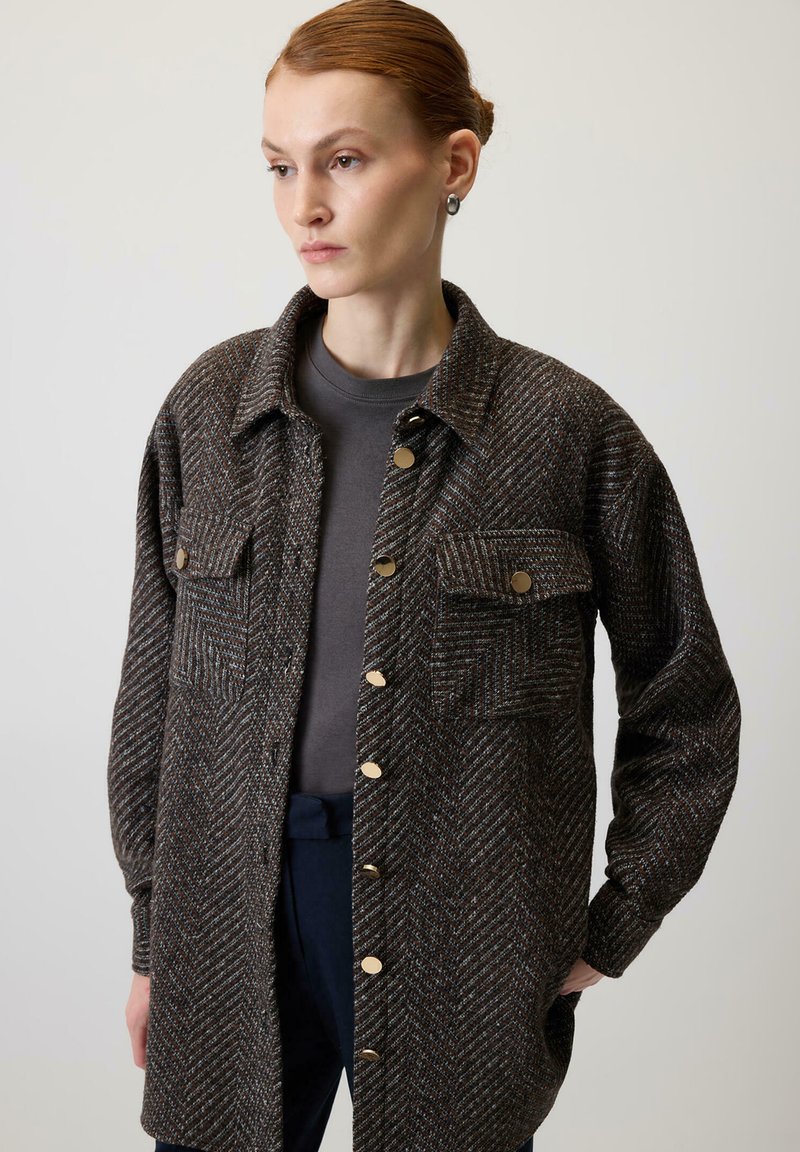 Brązowo-szara koszula typu shirt jacket o fakturze jodełki, z dwoma kieszeniami na piersi, złotymi guzikami i luźnym krojem, noszona na ciemnoszarym topie.