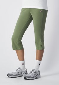 Zielone legginsy capri z subtelnym połyskiem, z rozcięciem po bokach i małą naszywką z logo na udzie. W połączeniu z szarymi sneakersami i białymi skarpetkami.