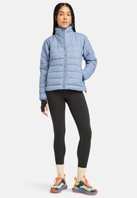 Ljusblå quiltad pufferjacka med horisontellt mönster, hög krage, dragkedja, i kombination med svarta leggings och färgglada sneakers.