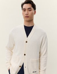 Cardigan crema con un tessuto a maglia testurizzato, scollatura rotonda, bottoni in legno e due tasche frontali. Presenta un delicato ricamo "les deux" sulla tasca.