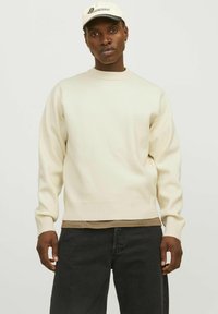 Pull crème avec des poignets et un ourlet côtelés, superposé à une chemise marron, associé à un jean noir, porté avec une casquette de couleur claire.