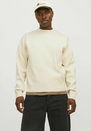 Crème trui met ribbels aan de mouwen en onderkant, gelaagd over een bruine shirt, gecombineerd met zwarte jeans, gedragen met een lichtgekleurde pet.