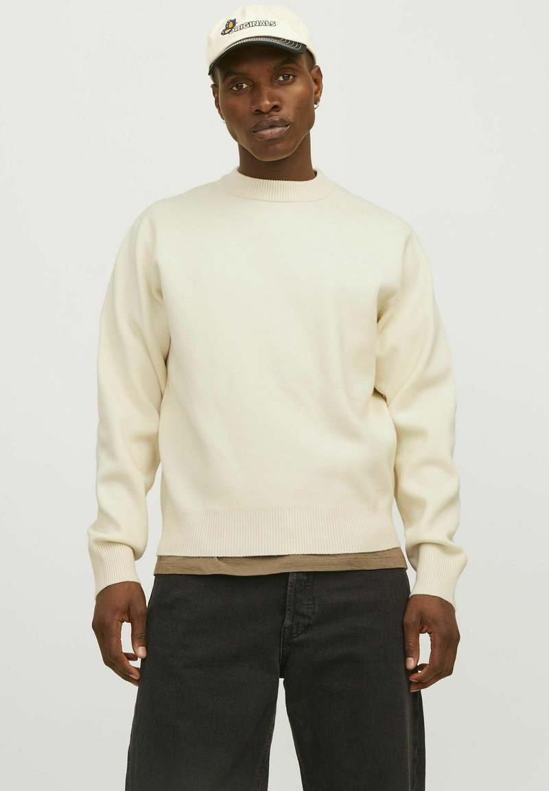 Pull crème avec des poignets et un ourlet côtelés, superposé à une chemise marron, associé à un jean noir, porté avec une casquette de couleur claire.