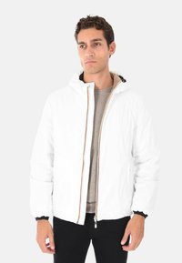 Uomo che indossa una giacca bianca con cappuccio sopra un maglione beige e pantaloni neri, in piedi contro uno sfondo chiaro e uniforme.