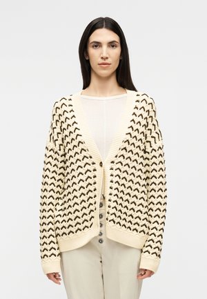 Γυναίκα που φοράει εκρού ζακέτα με μαύρο σχέδιο ζιγκ-ζαγκ, κουμπωμένη μπροστά, πάνω από διάφανο λευκό τοπ και μπεζ παντελόνι.