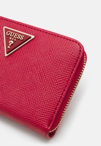 Rode portemonnee met textuur, ritssluiting en gouden hardware. Bevat een driehoekige logo-plaat met de tekst "GUESS USA". Compact ontwerp.