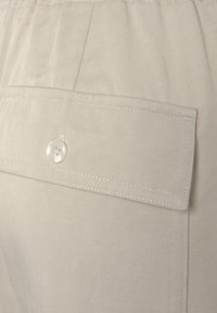Väriä beige olevaa puuvillakangasta teksturoidulla pinnalla, jossa on napilla suljettava takatasku ja hienovaraisia ompeludetaileja.