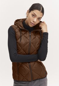 Gilet matelassé marron avec capuche, doté d'une fermeture éclair et de poches latérales, porté par-dessus un haut à manches longues noir. Texture lisse, design arrondi.