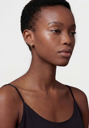 Nuori mustaihoinen nainen, jolla on lyhyet kiharat hiukset, hänellä on musta spaghettiolkaiminen paita ja pienet kultaiset renkaskorvakorut, katsoo oikealle.