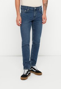 Blauwe denimjeans met een slim fit, voorzien van vijf zakken, een knoopsluiting en stikseldetails. Gecombineerd met zwart-witte sneakers.