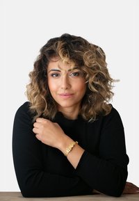Bracciale d'oro con design intricati indossato al polso, caratterizzato da una texture liscia e da accenti delicati. Il modello indossa abiti scuri e ha capelli ricci.