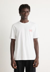 Ung mann med korte dreads iført en hvit T-skjorte med en liten rød "HUGO"-logo på brystet og svarte bukser, står foran en ensfarget bakgrunn.