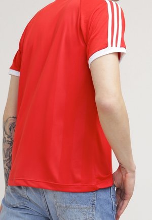 Personne portant une chemise rouge à manches courtes avec des rayures blanches sur les épaules et un jean bleu, montrant un tatouage sur l'avant-bras gauche.