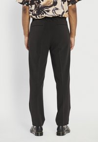 Pantalon noir à coupe ajustée, doté de deux poches arrière et d'une jambe droite, associé à des chaussures noires et une chemise à motifs.