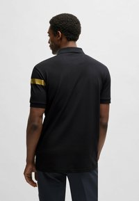 Sort polo shirt med krave, der har en gylden vandret stribe på venstre ærme. Lavet af glat stof, kortærmet, standard pasform.