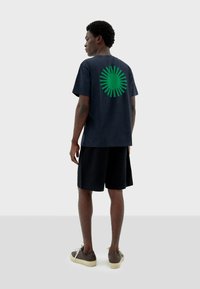 T-shirt bleu marine à manches courtes avec un motif de soleil vert au dos, associé à un short noir texturé et des chaussures brunes avec des lacets clairs.