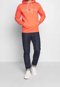 Felpa arancione con cappuccio e tasca frontale, testo del logo bianco e lacci. Abbinata a jeans scuri e sneakers bianche.