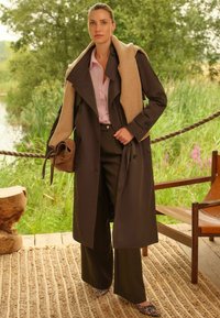 Dolga rjava plašč s bež ovratnikom, roza srajca, temni hlače in čevlji s tiskanjem leoparda. V roki drži zapestno torbico.