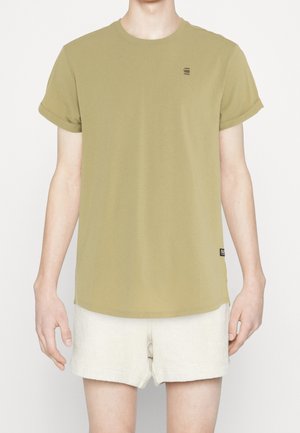 Person trägt ein olivgrünes kurzärmeliges T-Shirt mit hochgekrempelten Bündchen und hellbeige strukturierte Shorts vor einfarbigem Hintergrund.