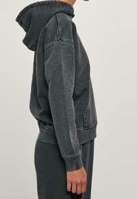 Persona che indossa una felpa con cappuccio grigio scuro e pantaloni abbinati, vista di lato su uno sfondo chiaro e uniforme.