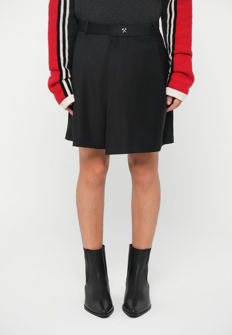 Falda negra con un diseño plisado, que presenta un cinturón con un pequeño acento metálico. Combinada con botines negros ajustados.