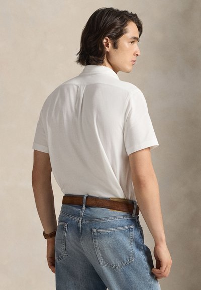 Camisa branca de manga curta com gola e costura nas costas, combinada com jeans de ganga azul clara e cinto de pele castanho.