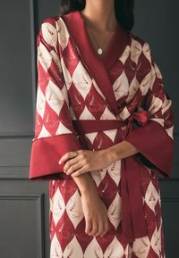 Punane ja kreemikas kimono-stiilis rüü, millel on luige muster, laiad varrukad, sidumisrihm ja sile tekstuur. Käe peal kuldsest ring.