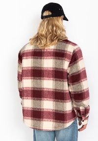 Persona con cabello largo rubio que lleva una gorra negra, una camisa de franela a cuadros en burdeos y beige, y jeans azul claro de espaldas sobre un fondo blanco.