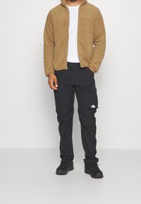 Brun fleece zip-jacka, vit t-shirt under, svarta cargo-byxor med logga, och svarta vandringsskor. Strukturerad tyg och funktionell design.