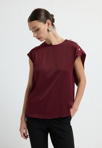 Blusa borgogna in tessuto liscio con maniche a cappuccio e bottoni decorativi argentati su una spalla. Abbinata a pantaloni neri.