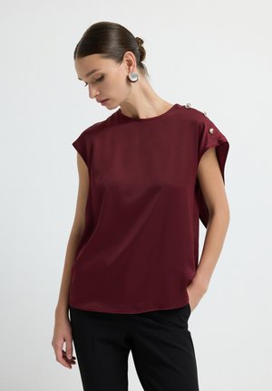 Bordeaux blouse van soepele stof met kapmouwen en decoratieve zilveren knopen op één schouder. Gecombineerd met zwarte broek.