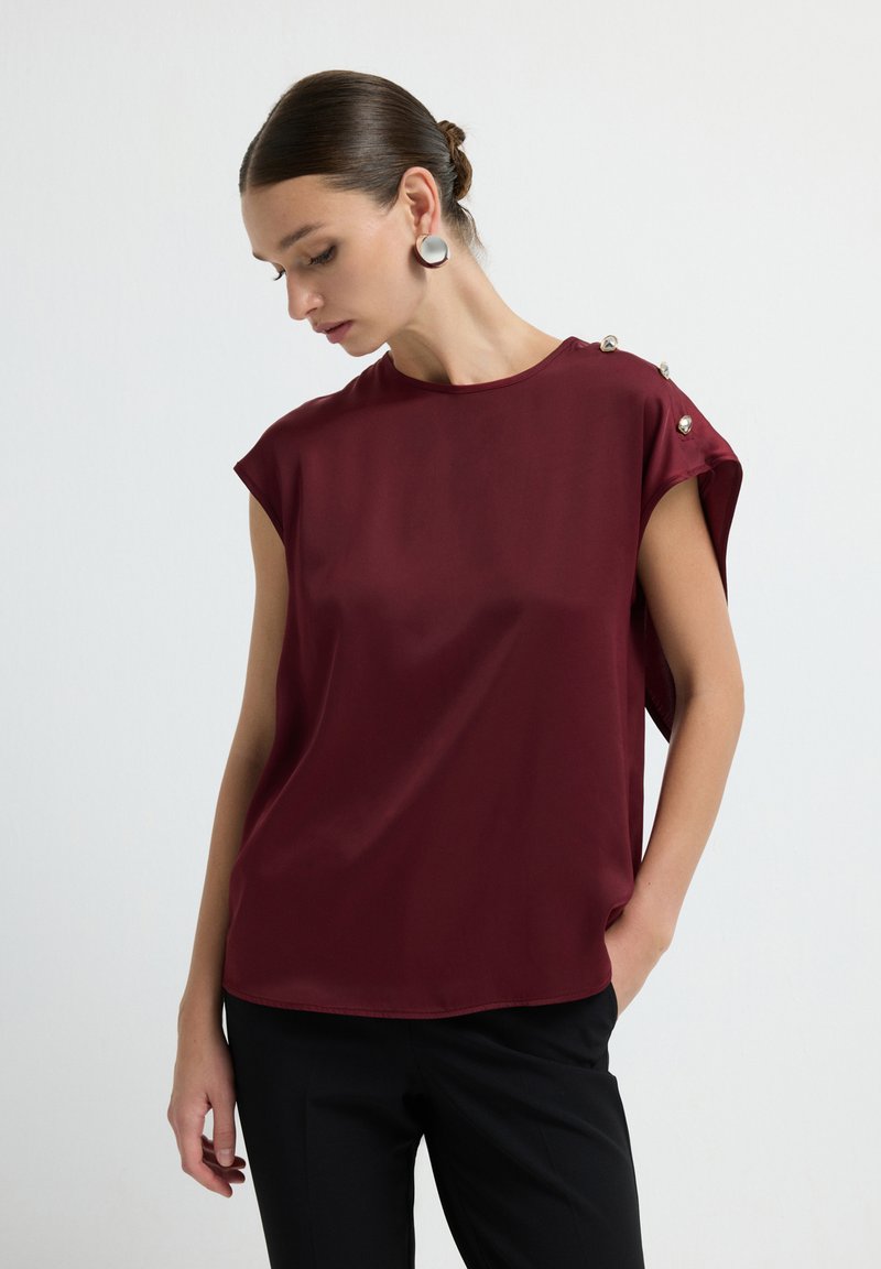 Blusa borgogna in tessuto liscio con maniche a cappuccio e bottoni decorativi argentati su una spalla. Abbinata a pantaloni neri.