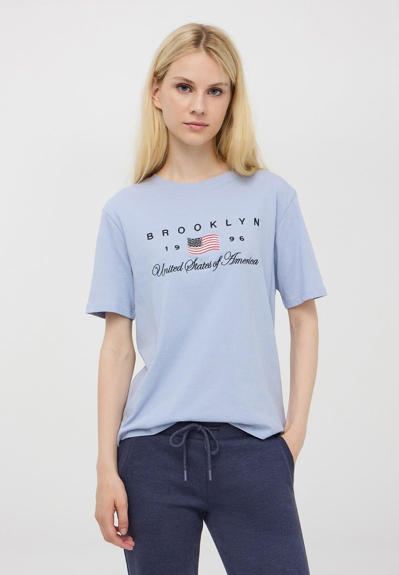T-shirt in cotone azzurro chiaro con maniche corte, presenta la scritta "BROOKLYN 1996" e una piccola grafica con la bandiera americana. Vestibilità morbida e casual.